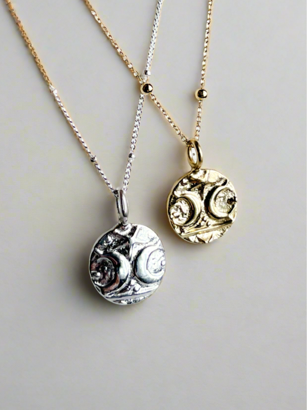 Two pendant necklaces, one with a silver coin-like pendant and the other with a gold vermeil coin-like pendant, both on chain necklaces.