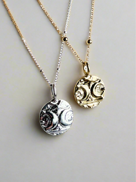 Two pendant necklaces, one with a silver coin-like pendant and the other with a gold vermeil coin-like pendant, both on chain necklaces.