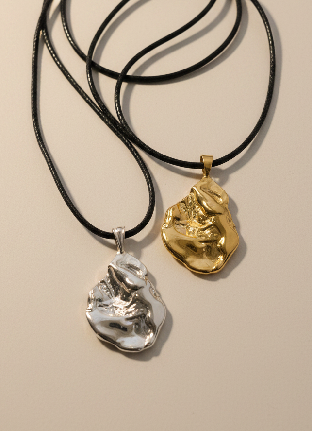 Two textured abstract pendant necklaces on black cords, one in polished silver and one in polished gold, displayed on a light gray surface.