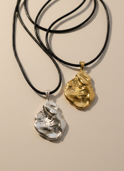 Two textured abstract pendant necklaces on black cords, one in polished silver and one in polished gold, displayed on a light gray surface.