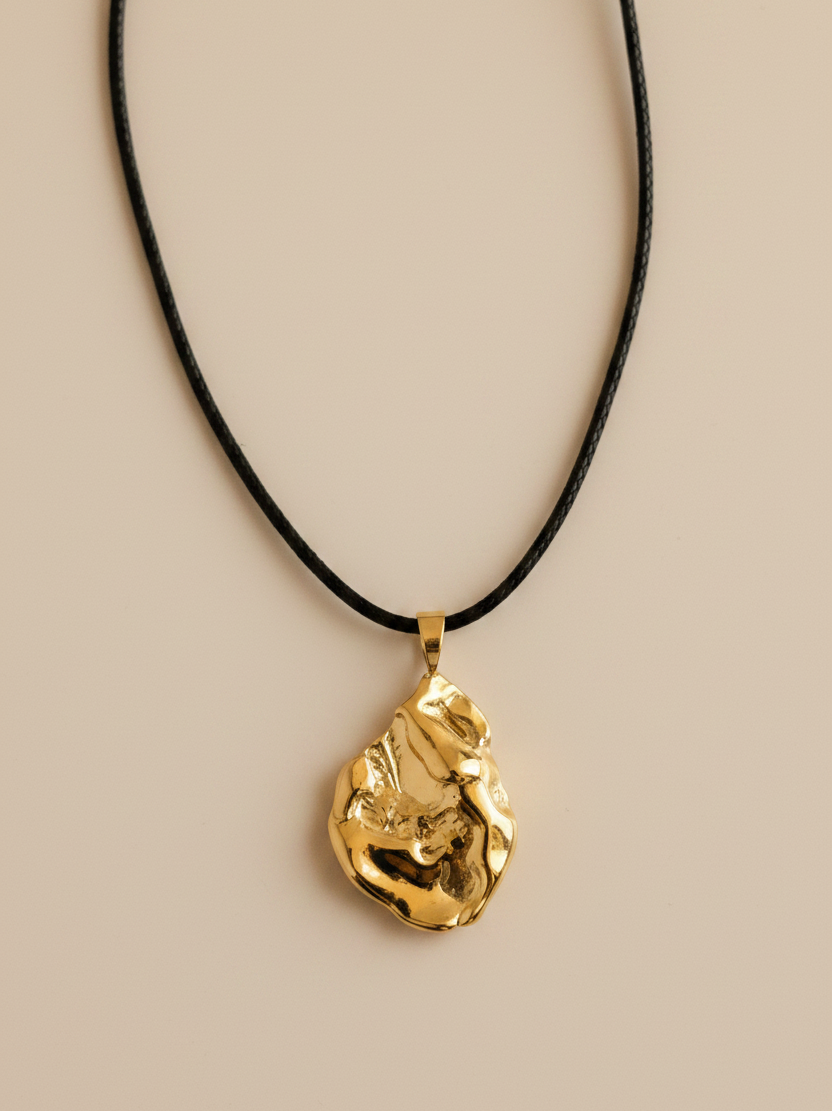 A gold pendant necklace with an organic, textured abstract shape hanging from a thin black cord, displayed on a light gray textured background.